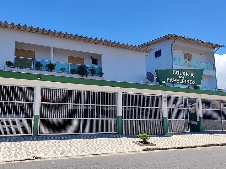 Fachada branca e verde da Colônia dos Papeleiros, com o portão da garagem e a placa com o nome